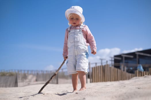 Little girl is sitting on sand and playing at sea beach in warm sunny summer day