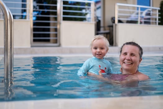 Mother and baby in outdoor swimming pool of tropical resort. Kid learning to swim. Mom and child playing in water. Family summer vacation in exotic destination. Active and healthy sport for kids.