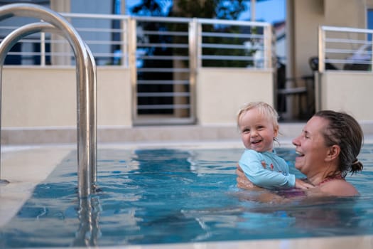 Mother and baby in outdoor swimming pool of tropical resort. Kid learning to swim. Mom and child playing in water. Family summer vacation in exotic destination. Active and healthy sport for kids.