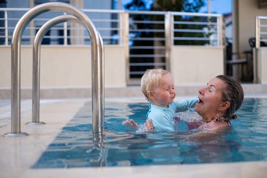 Mother and baby in outdoor swimming pool of tropical resort. Kid learning to swim. Mom and child playing in water. Family summer vacation in exotic destination. Active and healthy sport for kids.