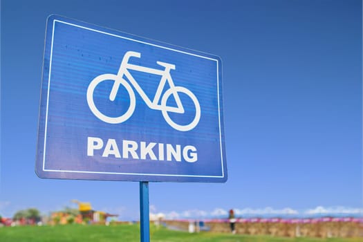A blue traffic sign featuring a bicycle symbol against a backdrop of the sky, grass, and plants. The font is bold and easy to read, emphasizing the importance of sharing the road with bicyclists