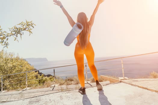 A woman wearing a bright orange sports outfit and carrying a yoga mat stands with her arms raised high on a mountain top, enjoying a breathtaking view.