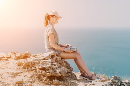 A young woman in a cap sits on a cliffside with a laptop, enjoying the view of the ocean on a bright sunny day.