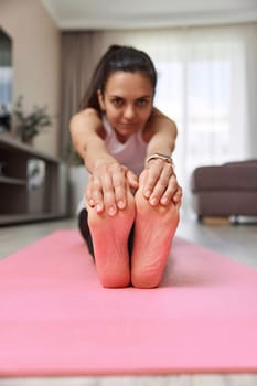 Sporty Caucasian woman worm up by stretching her body on pink exercise mat at home. focus on foot