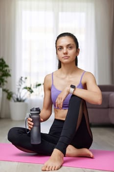 caucasian woman drinking water sitting on pink exercise mat at home