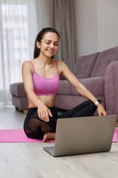 Happy Caucasian woman looking at laptop while sitting on pink fitness mat, ready for online training and workout at home. Online training.