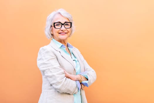 stylish mature entrepreneur woman smiling happy looking at camera leaning on an orange wall with arms crossed, concept of active lifestyle in adulthood, copyspace for text
