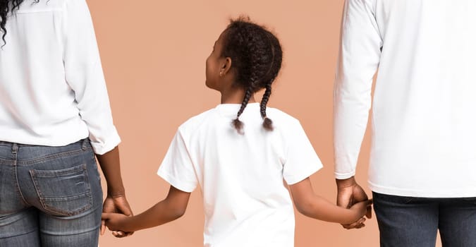 Family support. Little black girl with braids holding hands of her mom and dad while they walking together over yellow background, rear view