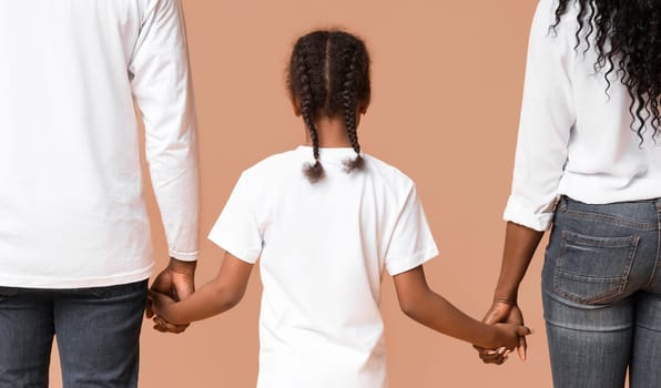 Adoption concept. Little afro girl holding hands of her adoptive parents, walking together over yellow background, rear view