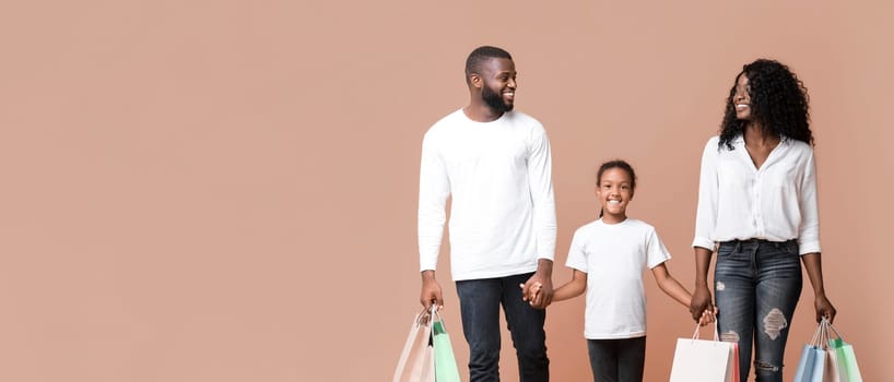 Family Shopping Concept. Happy African American parents and their daughter walking with shopper bags and holding hands in studio, yellow background.