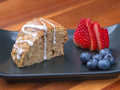 A close up of a slice of homemade zucchini bread with glaze, blueberries, and a sliced strawberry on a plate.