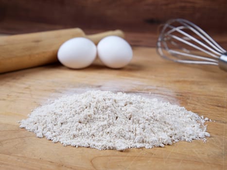 A close up of a pile of flour along with eggs and a rolling pin on a wooden cutting board.