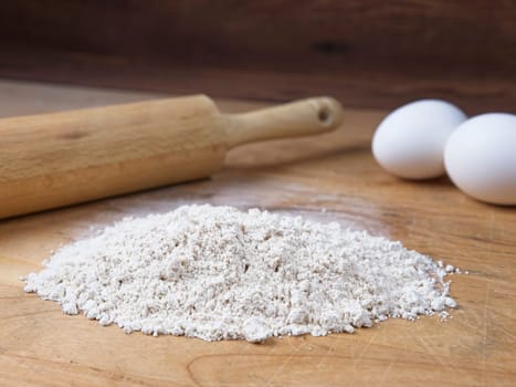 A close up of a pile of flour along with eggs and a rolling pin on a wooden cutting board.