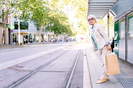 smiling mature woman standing with shopping bags waiting for public transport, concept of sustainable mobility and urban lifestyle, copyspace for text