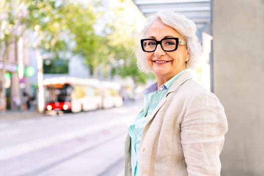 portrait of a smiling mature woman looking at camera while waiting for public transport, concept of sustainable mobility and urban lifestyle, copyspace for text