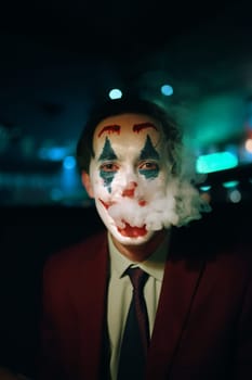 Person in suit and tie with the Joker makeup puffs smoke out of his mouth. Halloween party in nightclub with decorations and scary costumes. Autumn holiday. All Saints' Night.
