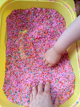 Mammy mother and baby play with touch dyed coloured rice in sensory bin. Sensory play. High quality photo. Sensory bin idea for child development