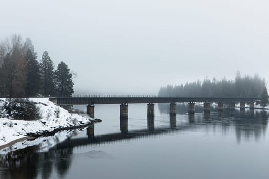 A railroad bridge crosses over the calm Clark Fork River on a foggy winter day in December in north Idaho.