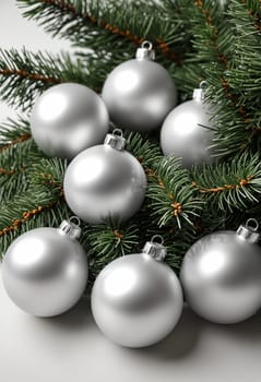 A close-up image of silver Christmas ornaments arranged within a cluster of evergreen branches. The ornaments are round and shiny, reflecting the light in a dazzling manner. The green branches provide a lush backdrop for the festive decorations.