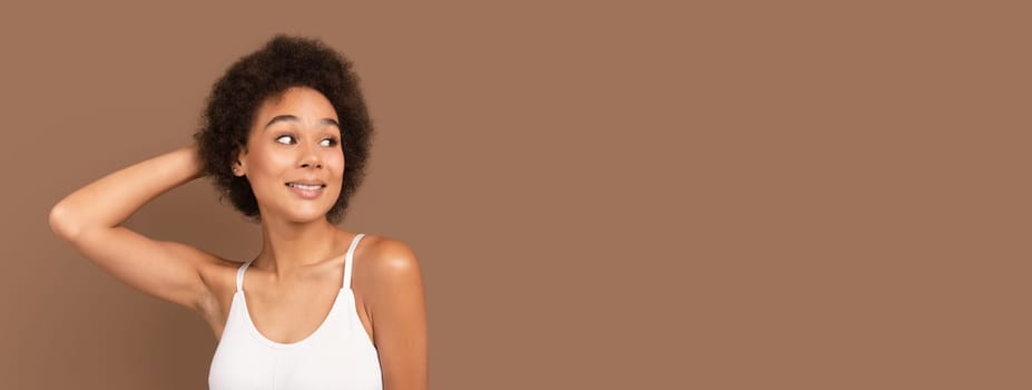 Cheerful millennial african american curly woman in white top with perfect skin looks at free space, isolated on brown background, panorama. Lady enjoy spare time, cosmetology, beauty care at home