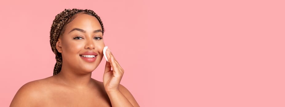 Facial skincare concept. Happy black chubby woman using cotton pad, caring for smooth skin, looking and smiling at camera, posing over pink studio background. Beauty and cosmetics concept, copy space
