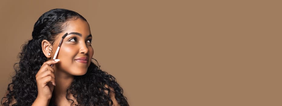 Beautiful brazilian body positive woman brushing eyebrows holding brush and looking aside at free space, enjoying beauty routine, brown studio background, copy space