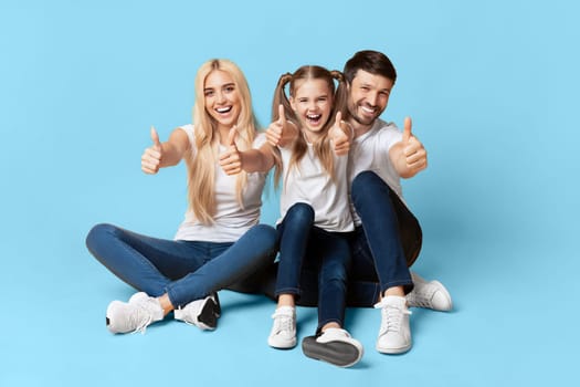 A family of three, a mother, father, and their young daughter, are sitting on a light blue background. They are all smiling and giving the thumbs up.