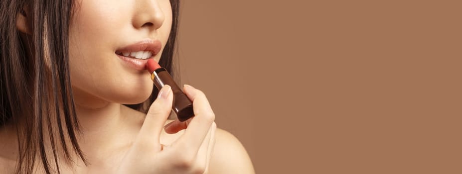 Cropped of unrecognizable asian lady using colorful lipstick over brown studio background, copy space. Lipstick, lipbalm, beauty products for lips, makeup concept