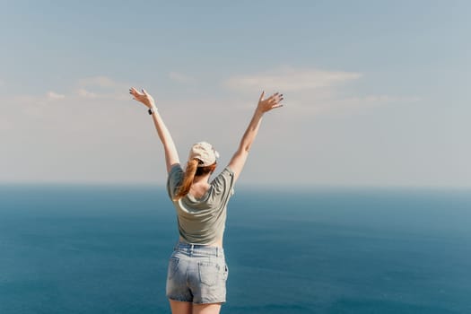 A young woman in a green shirt and denim shorts stands with her arms raised in celebration, enjoying the view of the blue ocean and the bright sky.