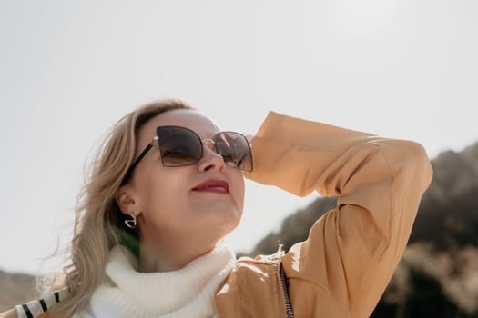 A woman with blonde hair and sunglasses looks up at the sky on a bright day, her yellow jacket and white sweater standing out against the blue sky.