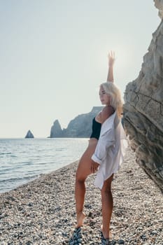 A woman in a black bathing suit and white shirt stands on a pebbled beach, arms outstretched, gazing towards the sea.