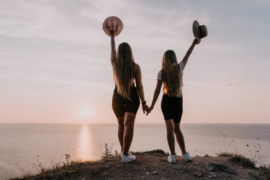 Two women in black shorts and white shirts stand on a cliffside holding hands, looking towards a setting sun over the ocean.