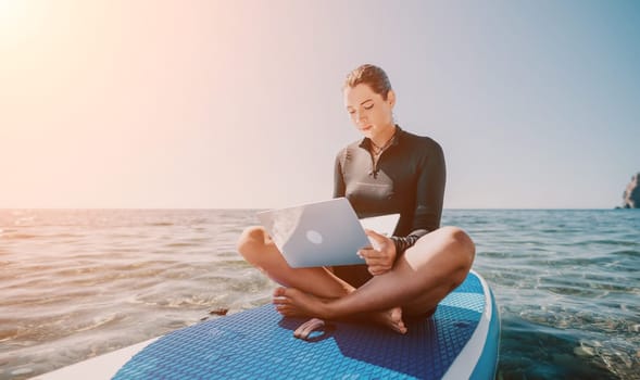 Digital nomad, woman in the hat, a business woman with a laptop sits on the rocks by the sea during sunset, makes a business transaction online from a distance. Freelance, remote work on vacation.