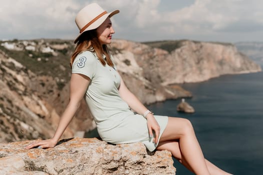 A woman wearing a light green dress and a straw hat sits on a rock cliff and looks out at the ocean.