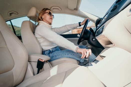 A woman in sunglasses wearing a white sweater and blue jeans sits in the driver's seat of a car, putting the car in gear.