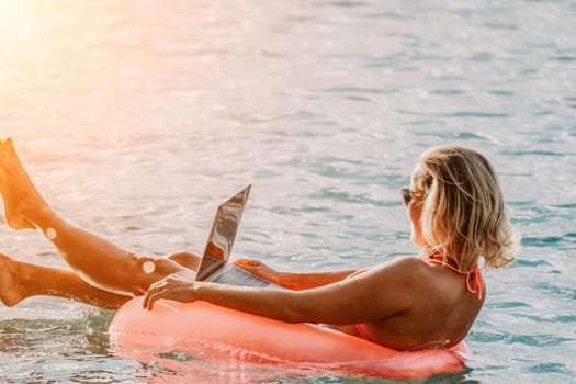 Woman works on laptop in sea. Freelancer, young blond woman in sunglases floating on an inflatable big pink donut with a laptop in the sea at sunset. Freelance, travel and holidays concept