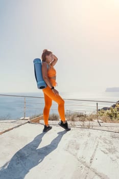 A woman in a bright orange yoga outfit stands on a concrete platform overlooking the sea, carrying a blue yoga mat. She gazes out at the horizon with a confident and serene expression, the sun casting a warm glow on the scene.