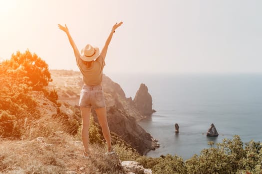 A woman with her arms raised in the air, enjoying a scenic view of the ocean from a cliff on a bright sunny day.