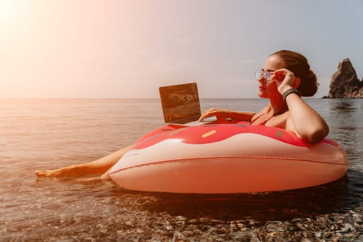 Woman freelancer works on laptop swimming in sea on pink inflatable ring. Pretty lady typing on computer while floating in the sea on inflatable donut at sunset. Freelance, remote work on vacation