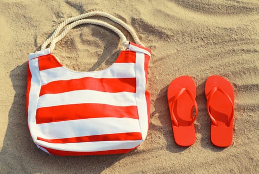 Summer vacation, accessories - red striped bag with flip flops lying on the sand on the beach, top view