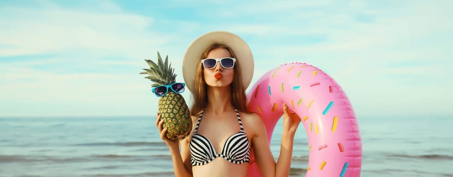 Summer vacation, tourism, happy young woman with swimming inflatable ring, pineapple fruits, tourist hat and glasses on the beach against sea background