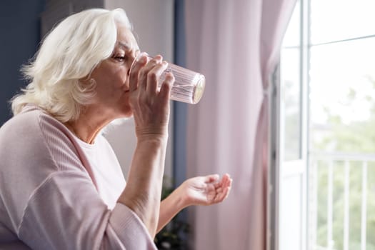 Senior woman takes pill with glass of water in hand. Stressed female drinking sedated antidepressant meds. Woman feels depressed, taking drugs.