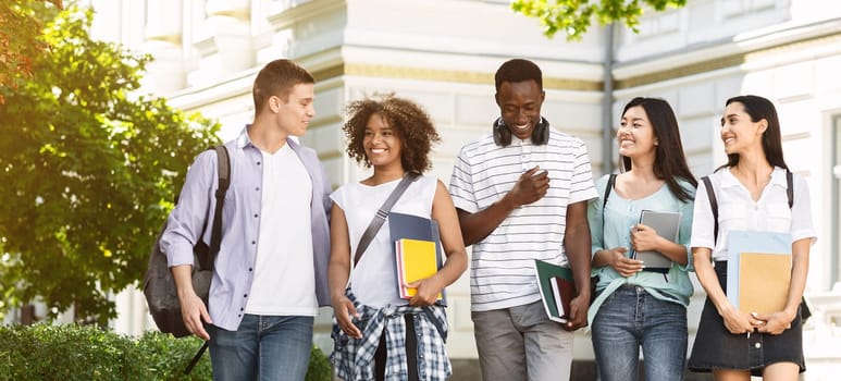Group of multiethnic students walking together outdoors in college campus, holding books and notepads and laughing, copy space