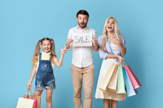 A family of three stands in front of a blue background, excitedly holding shopping bags and a sign that says Sale. The father holds the sign, the mother holds multiple shopping bags