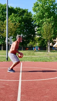 An elderly old Italian man plays basketball on a sports court on a summer day. Vertical