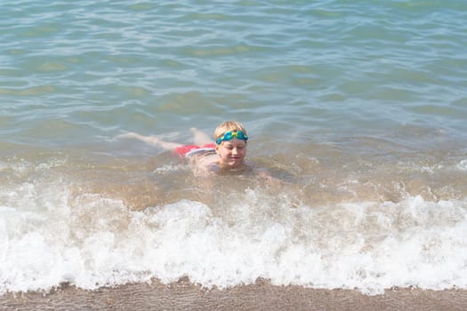 boy 8 years old in swimming goggles lies in the surf on the sandy beach of the blue sea, loves to swim during the summer holidays in Turkey, refreshing swim on a hot sunny day