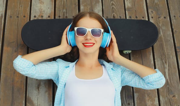 Happy young woman listening to music with headphones lying on a skateboard, modern teenager girl resting in city park