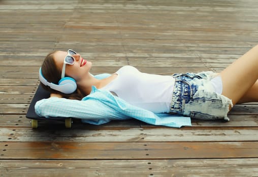 Happy young woman listening to music with headphones lying on a skateboard, modern teenager girl resting in city park