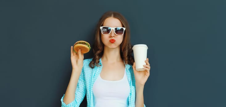 Portrait of happy cheerful young woman eating burger fast food holding cup of coffee, teenage girl having fun isolated on black wall studio background