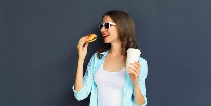 Portrait of happy cheerful young woman eating burger fast food holding cup of coffee, teenage girl having fun isolated on black wall studio background